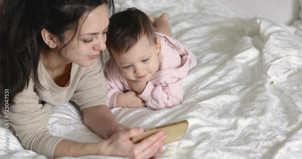 Mother and child daughter playing with smartphone while sitting on bed in bedroom at home Mother and child daughter playing with smartphone while sitting on bed in bedroom at home