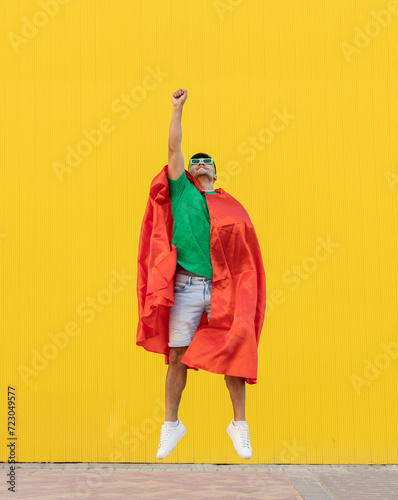 Young man wearing red cape jumping in front of yellow wall photo