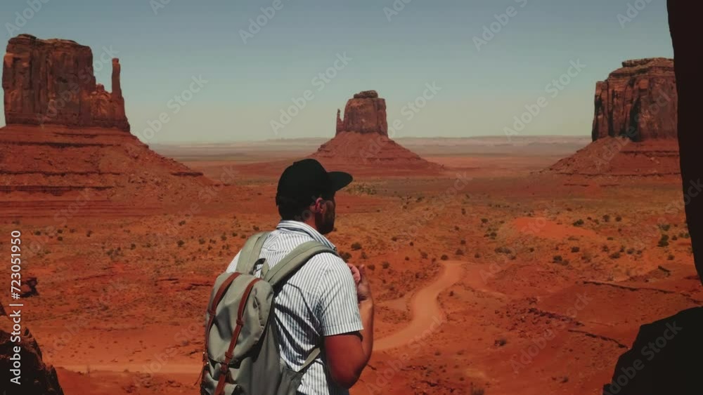 Back view young adult tourist man with backpack watching epic desert view, checking the phone over Monuments Valley. Back view young adult tourist man with backpack watching epic desert view, checking the phone over Monuments Valley.