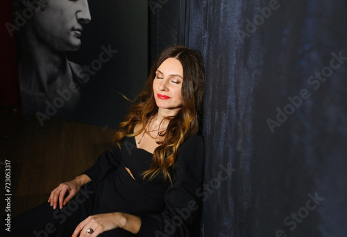 Relaxed girl closing her eyes while sitting indoors in a black romper. Background - blue textured wall, painting. Beauty concept