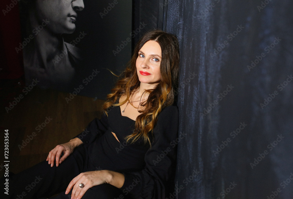 Stunning portrait of a girl looking at the camera while sitting indoors in a black romper. Background - blue textured wall, painting. Fashion and beauty concept