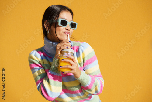Young woman drinking orange juice standing in front of yellow wall photo