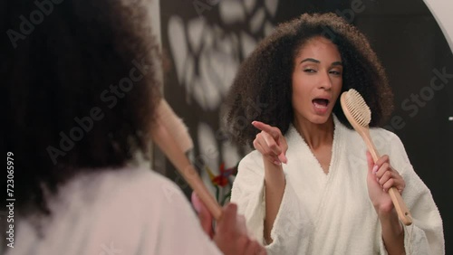 Happy active African American woman girl with curly hair in bathroom pointing in mirror reflection singing song in bath brush sing dancing having fun in morning beauty routine enjoy pampering hygiene photo