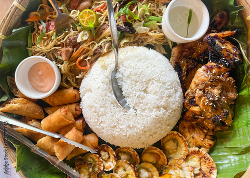 Filipino food on a plate with a banana leaf base. Rice, chicken, lumpia, pansit