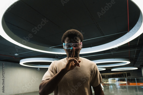 Young man with futuristic cyber glasses under modern ring lamp putting forefinger on lips photo