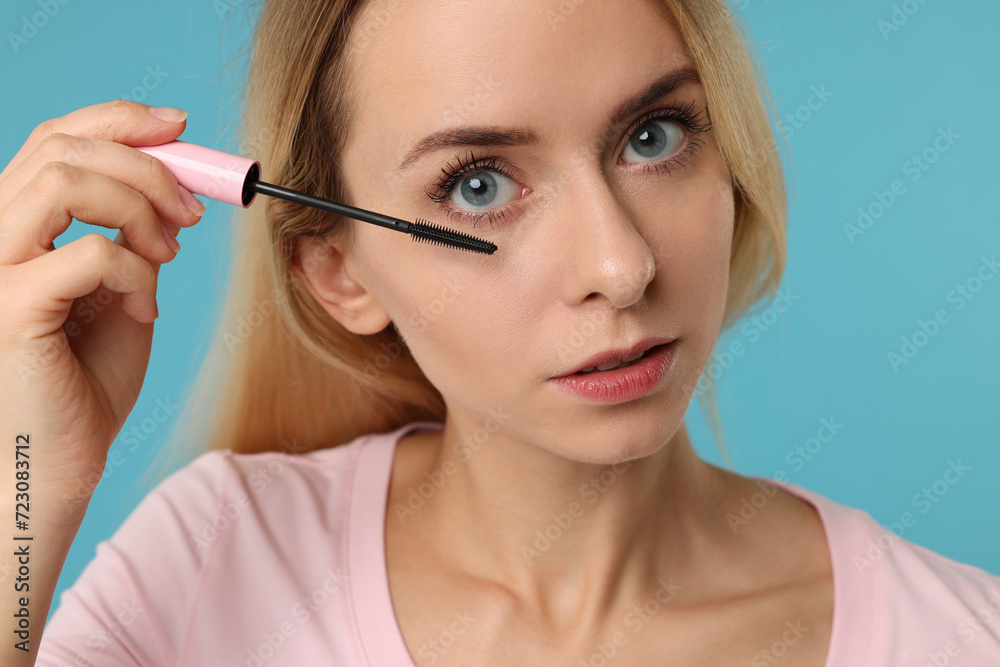 Beautiful woman applying mascara on light blue background, closeup