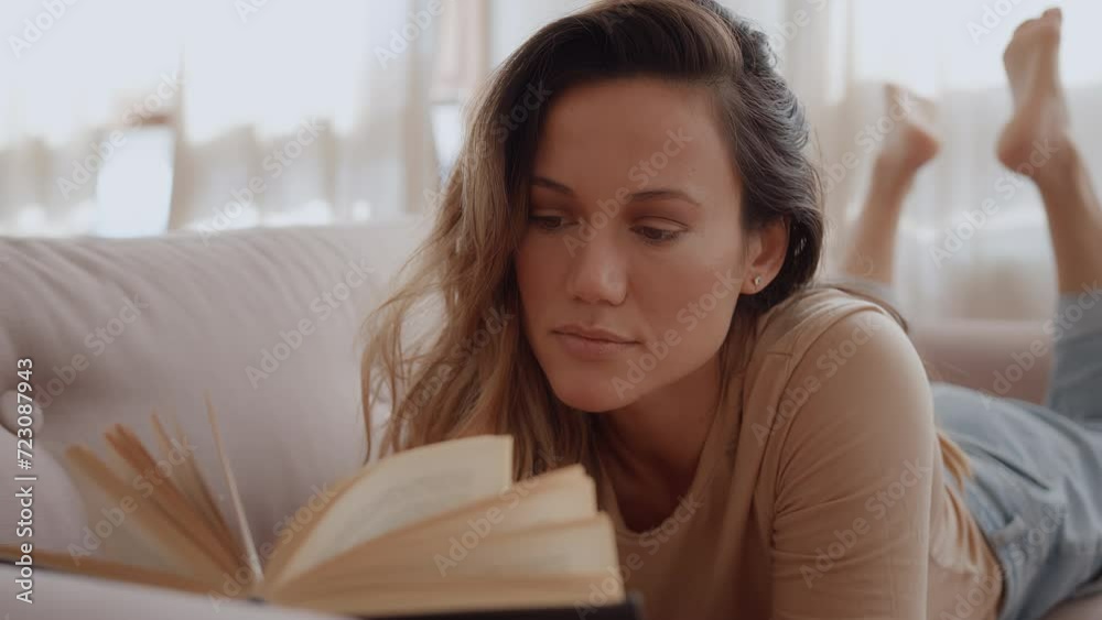 young contented woman lay on couch at home reading a book, looks pleased and cheerful due to story. female adult student learn textbook manual to prepare for exam