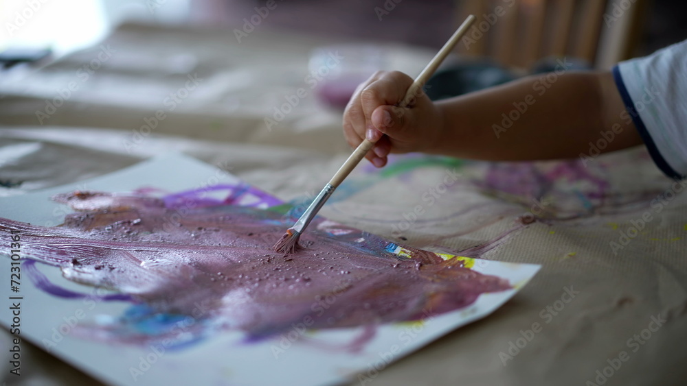 Obraz premium Closeup child hand holding paintbrush painting watercolor abstraction on paper, 3 year old kid engaged with artistic work at home