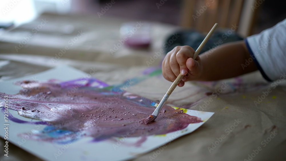 Obraz premium Closeup child hand holding paintbrush painting watercolor abstraction on paper, 3 year old kid engaged with artistic work at home
