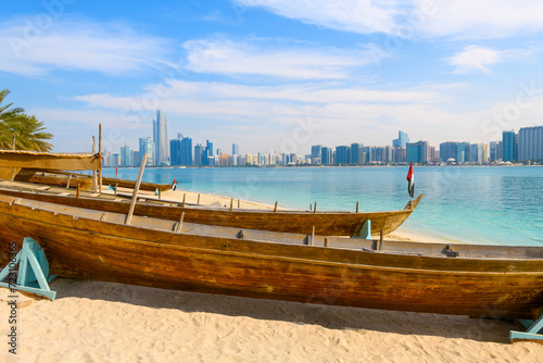 Traditional Arabic boats docked on a sandy beach at Marina Mall Island, with the waterfront corniche and skyline across the sea in Abu Dhabi, United Arab Emirates. photo