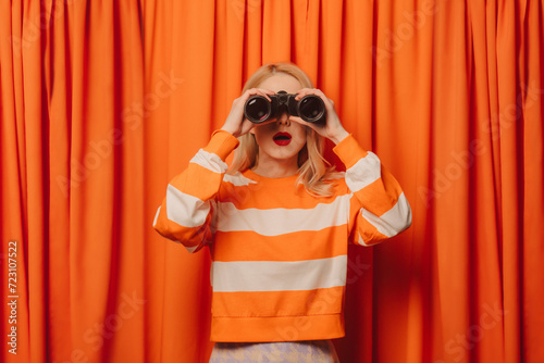 Woman looking through binoculars standing in front of orange curtain photo