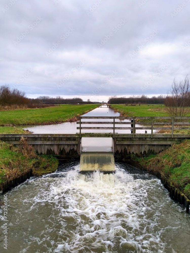 A floodgate between two canals with a small artificial waterfall and a wooden railing. Dutch landscape. 
