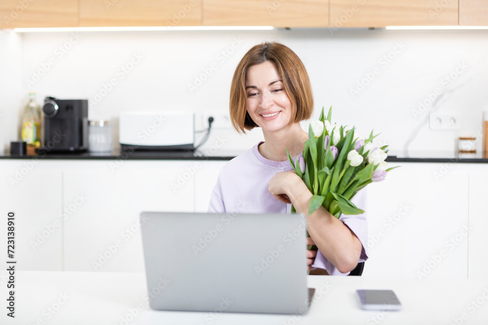 Young happy woman with bouquet of flowers sitting at the table and talking on video call on laptop. Online dating and celebrating holidays far from each other concept Young happy woman with bouquet of flowers sitting at the table and talking on video call on laptop. Online dating and celebrating holidays far from each other concept