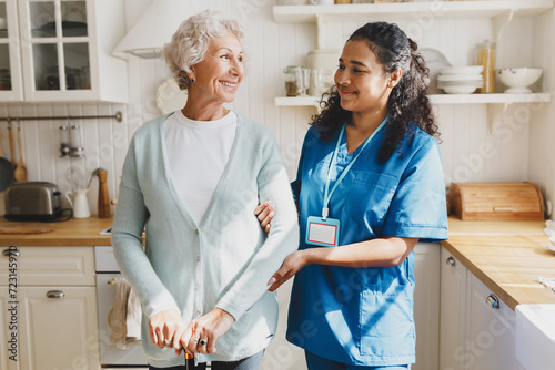 Medical assistance at home. Pretty african american female volunteer in blue uniform helping senior caucasian woman walk with walking stick, holding her by hand, looking at each other with smile photo