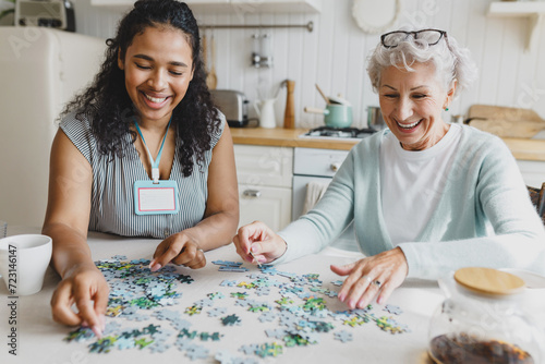 Portrait of african american female social worker volunteer playing puzzle game with cheerful senior lady, having fun, laughing searching for right piece sitting together at kitchen table photo