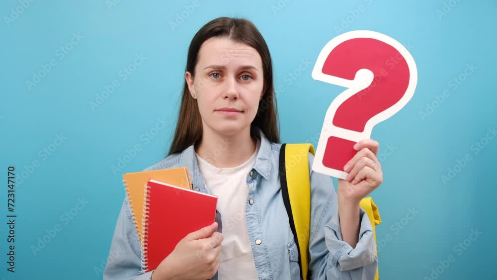 Portrait of pensive young woman student holding red question marks and books, wears shirt and yellow backpack, posing isolated over blue studio background wall. Education in university college concept Portrait of pensive young woman student holding red question marks and books, wears shirt and yellow backpack, posing isolated over blue studio background wall. Education in university college concept