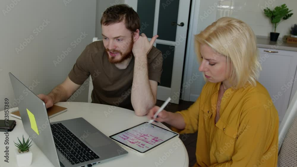 Young blond woman drawing with stylus pen in tablet while sitting at the table near young bearded man working on laptop in the kitchen. Young blond woman drawing with stylus pen in tablet while sitting at the table near young bearded man working on laptop in the kitchen.