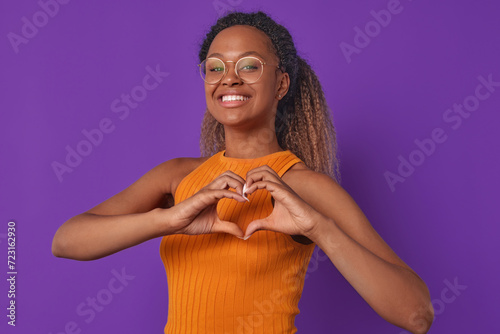 Young cheerful beautiful African American woman in glasses shows heart shape from her fingers confessing love or romantic feelings causing passion and sympathy stands in purple studio. Valentine day.