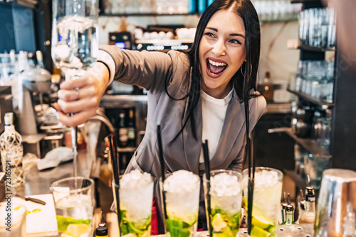 Woman barman preparing alcoholic cocktail and pouring it into glass with crushed ice, mint and lime at bar - Barlady smiling while pouring rum to prepare mojito at pub - Female work in the night club photo
