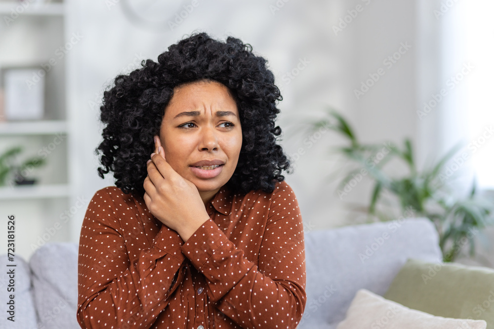 Close-up photo of a young upset African-American woman sitting on the couch at home and suffering from ear pain, holding her head with her hand Close-up photo of a young upset African-American woman sitting on the couch at home and suffering from ear pain, holding her head with her hand