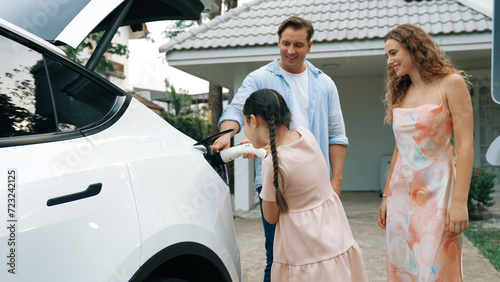 Happy little young girl learn about eco-friendly and energy sustainability as she help her family recharge electric vehicle from home EV charging station. EV car and modern family concept. Synchronos photo