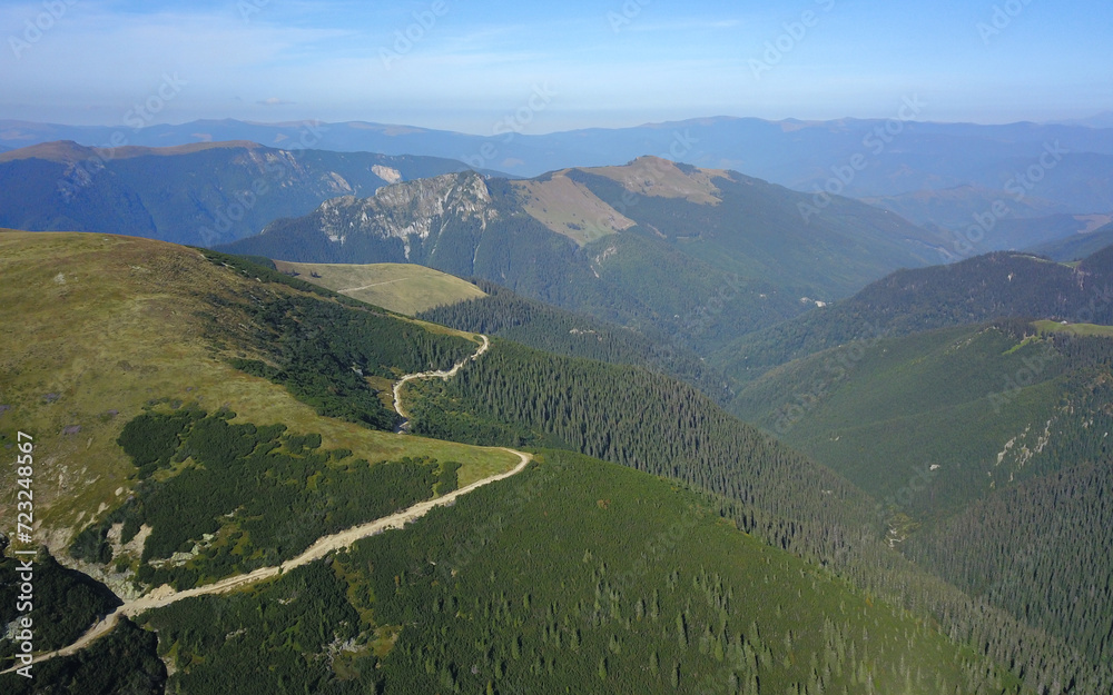 Aerial drone view of a gravel road winding at a high altitude along mountaintops, alpine pastures and coniferous forests beneath. Capatanii Mountains, Romania.