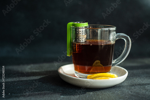 A warm cup of tea with a green tea infuser and lemon slices on a saucer, placed on a dark textured table photo