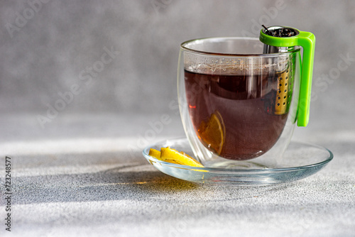 A transparent cup of hot tea with a slice of lemon on a saucer and a green tea infuser hanging on the side photo