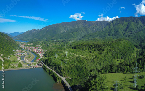 Aerial drone view above a rural settlement at the feet of Carpathian Mountains. An asphalt road winds over a lake, and a dam, over concrete bridges. Electricity pylons transfer power over the hills. 