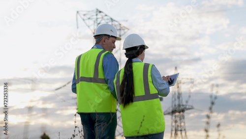 Engineer and trainee with tablet review power transmission lines walking along substation area at sunset. Electricians with tablet plans repair of power plant and transmission lines. Electricians work