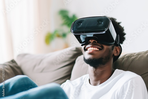 A young African-American man smiles while wearing virtual reality goggles on his living room couch. © aguadeluna