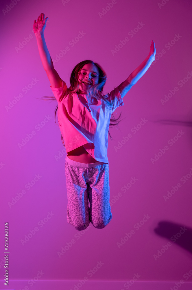 Portrait of cute smiling girl jumping with hands up on pink background with blue neon light. Party concept. Different emotions. Happiness, childhood, holidays, women's day, lifestyle. Copy space. 