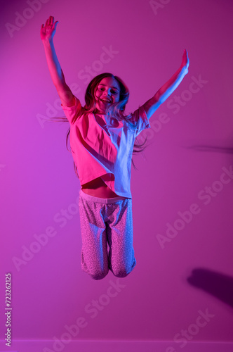 Portrait of cute smiling girl jumping with hands up on pink background with blue neon light. Party concept. Different emotions. Happiness, childhood, holidays, women's day, lifestyle. Copy space. 