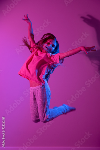 Portrait of cute smiling girl jumping with hands up on pink background with blue neon light. Party concept. Different emotions. Happiness, childhood, holidays, women's day, lifestyle. Copy space. 