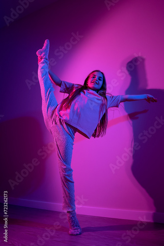 Cute smiling flexible girl with one leg up doing gymnastic exercise on pink background with blue neon light. Party concept. Different emotions. Happiness, childhood, holidays, women's day, lifestyle. 
