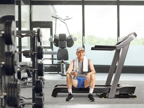 Elderly man sitting on a treadmill at a gym with a bottle of water