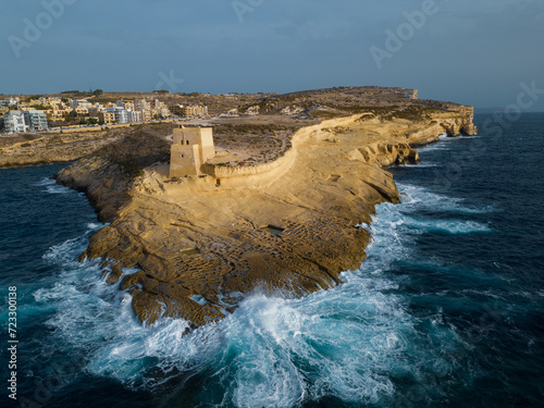 Aerial view of spectacular Gozo island coastline with cliffs. In front rough waves against the rock with historic Xlendi Tower on top. At the back of the bay Xlendi town with tourist accommodations. photo