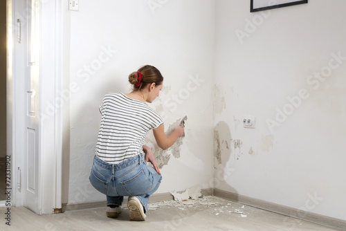 Saltpeter on the wall problem. Woman is using a scraper to scrape and remove all loose paint and plaster that is in poor condition, until a firm surface is achieved.  photo