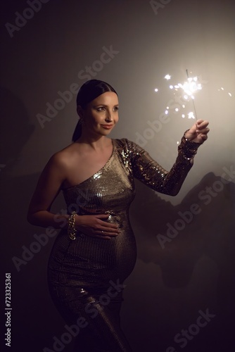 pregnant woman in a gold dress holds a sparkler in her hands on New Year's Eve on the background of a white wall in an apartment.