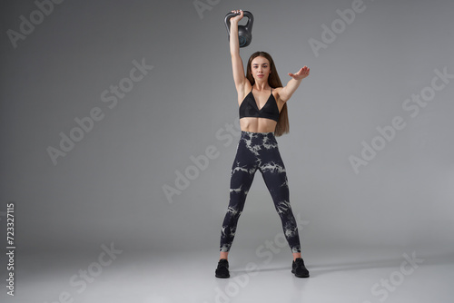 Attractive Caucasian woman in sportswear, lifting heavy kettle bell in studio. Front view of strong fair-haired female with raised arm, lifting weight and smiling, isolated on grey. Sport concept.