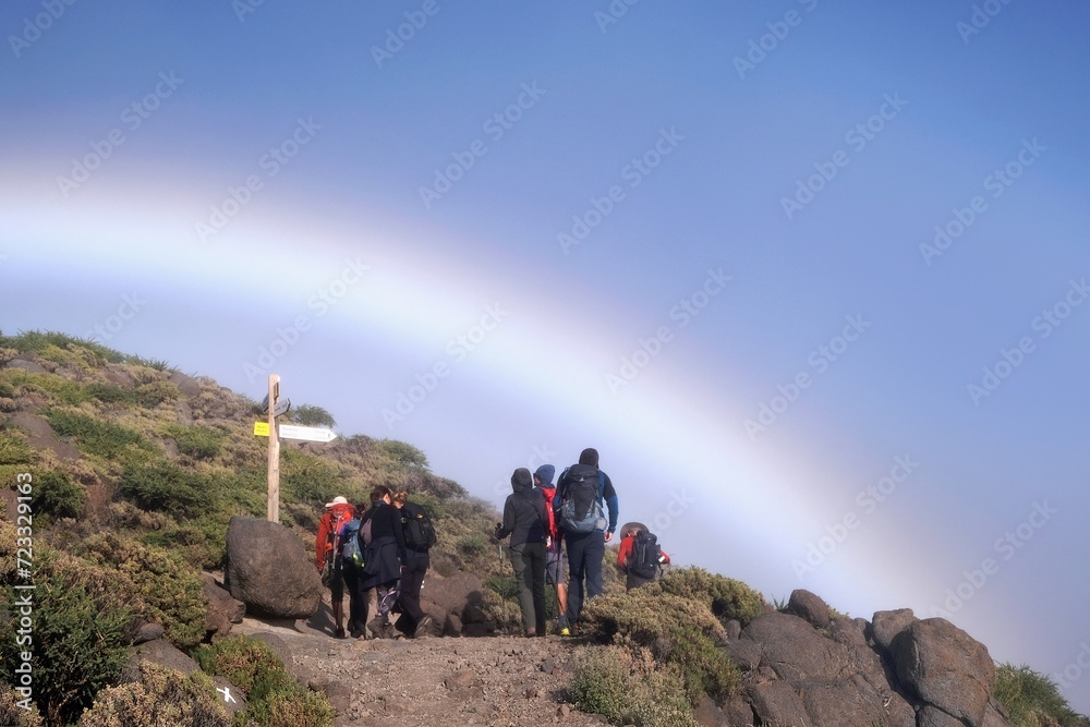 Group of hiking tourist by signpost to peak Pico de la Nieve in National Park Caldera de Taburiente. Rainbow on blue sky on background. La Palma, Canary Islands, Spain