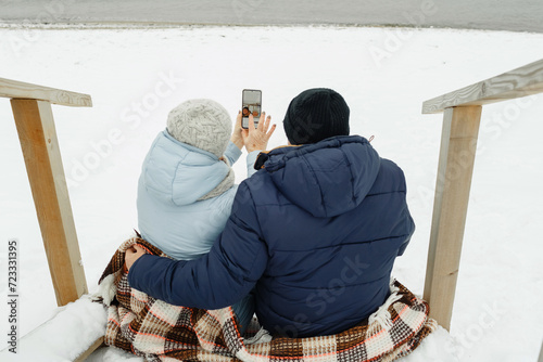 An elderly couple sitting on the stairs by the sea in winter, taking a selfie.