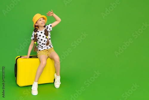 A young girl with a yellow suitcase is going on a trip. A teenage girl in a summer hat and shorts sits on her luggage and looks into the distance smiling broadly. Green isolated background. photo