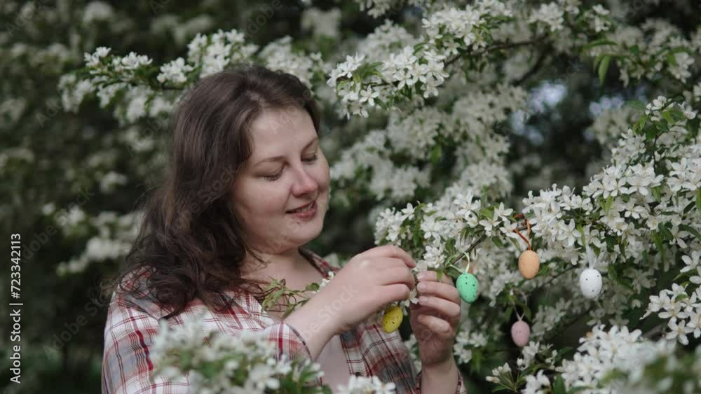 Caucasian woman decorates blooming spring tree with decorative egg. Portrait of housewife prepares fruit tree in home garden for Easter. Symbolic Easter decorations. Caucasian woman decorates blooming spring tree with decorative egg. Portrait of housewife prepares fruit tree in home garden for Easter. Symbolic Easter decorations.