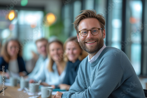 Portrait of a handsome young man in glasses standing in a creative office with his colleagues in the background