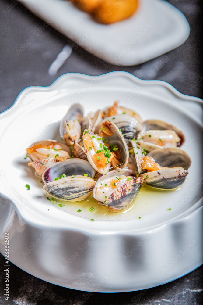 Almejas a la marinera, decoradas con cebollino y salsa de vino albariño en un plato blanco sobre una mesa de marmol negro acompañadas de coquetas de marisco Almejas a la marinera, decoradas con cebollino y salsa de vino albariño en un plato blanco sobre una mesa de marmol negro acompañadas de coquetas de marisco