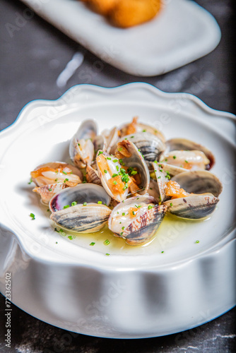 Almejas a la marinera, decoradas con cebollino y salsa de vino albariño en un plato blanco sobre una mesa de marmol negro acompañadas de coquetas de marisco