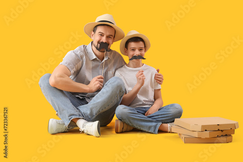 Portrait of father and his little son with paper mustache and pizza on yellow background