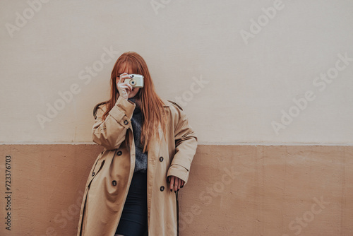 A trendy lady is taking pictures with her camera while leaning on a wall. photo