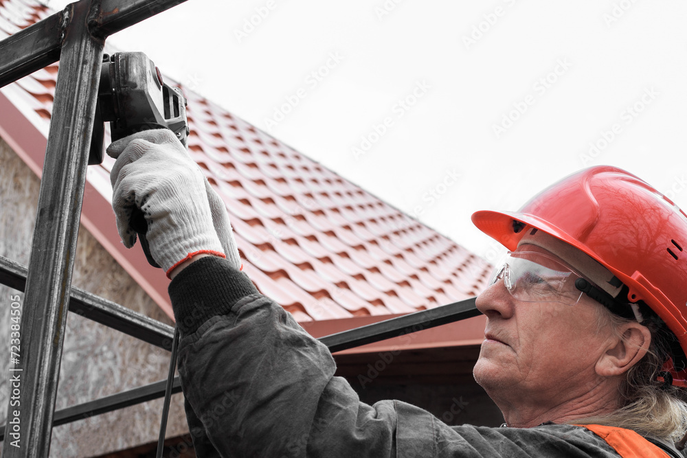 A worker in a helmet and safety glasses works with a grinder with a profile pipe. Installation and construction